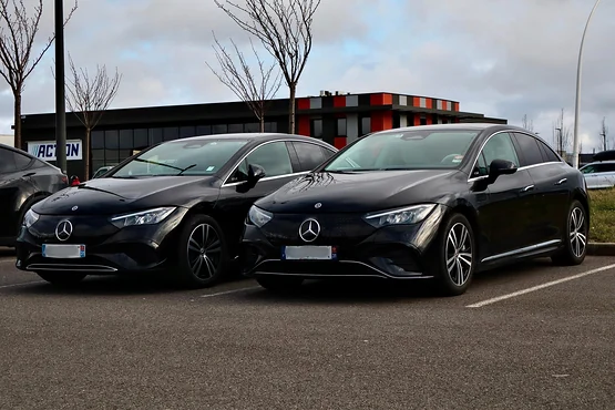 Deux berlines noires stationnées côte à côte sur un parking, devant un bâtiment et des arbres sans feuilles.