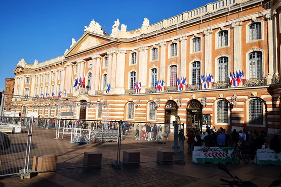 Façade du Capitole de Toulouse photographiée depuis la place, avec les drapeaux au balcon et les terrasses au premier plan.