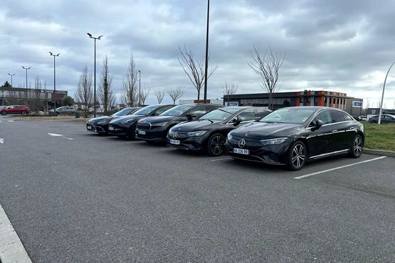 Plusieurs berlines noires alignées sur un parking en plein air, sous un ciel nuageux.